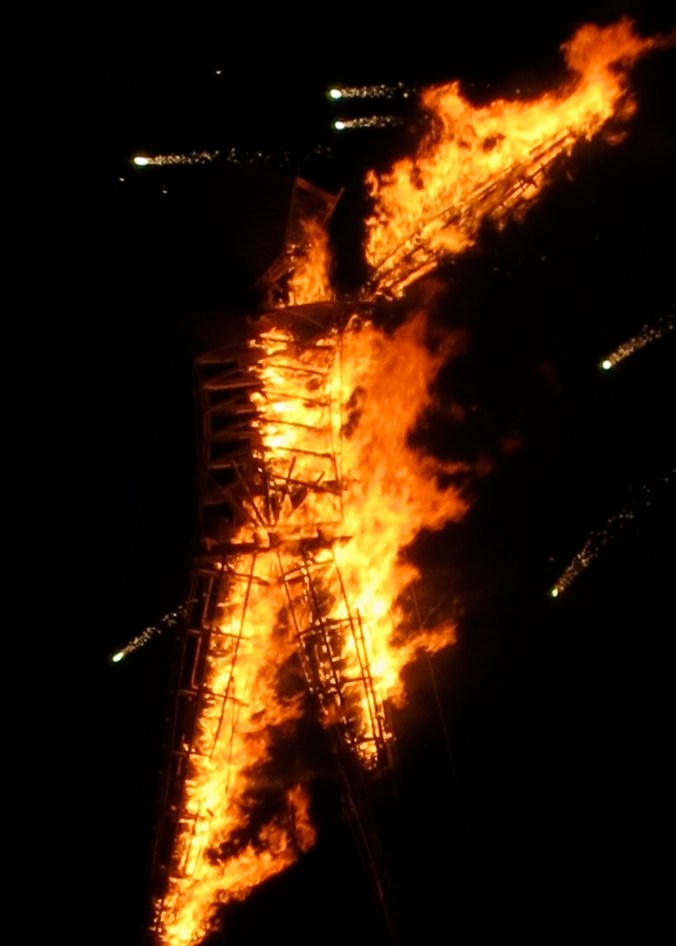 The Man burns at Burning Man while fireworks shoot across the sky.