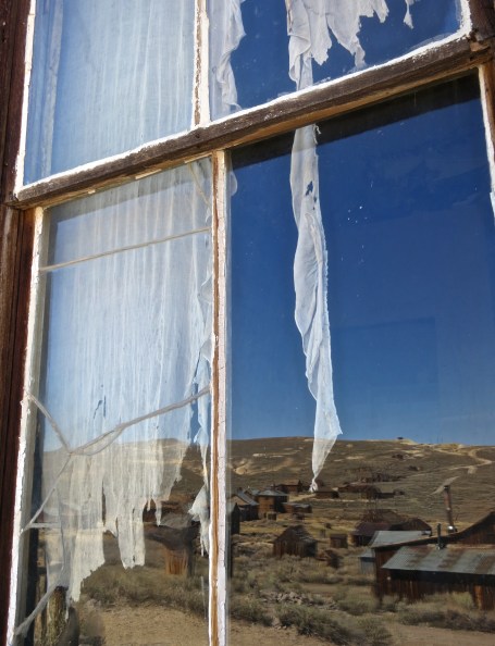 Tattered curtains, a cracked window, and a reflection of weather warn buildings capture the essence of the Old West ghost town of Bodie, California.